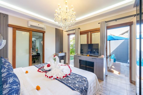 Luxurious Bali villa bedroom with king bed adorned in rose petals and swan towels, crystal chandelier, flat-screen TV, and private infinity pool view.